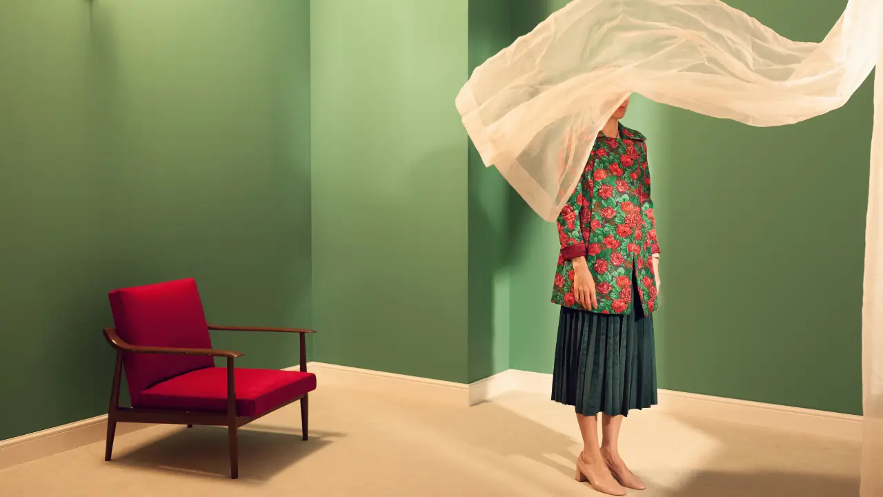 A woman wearing a floral coat stands in a room with a voile curtain blowing across her face.