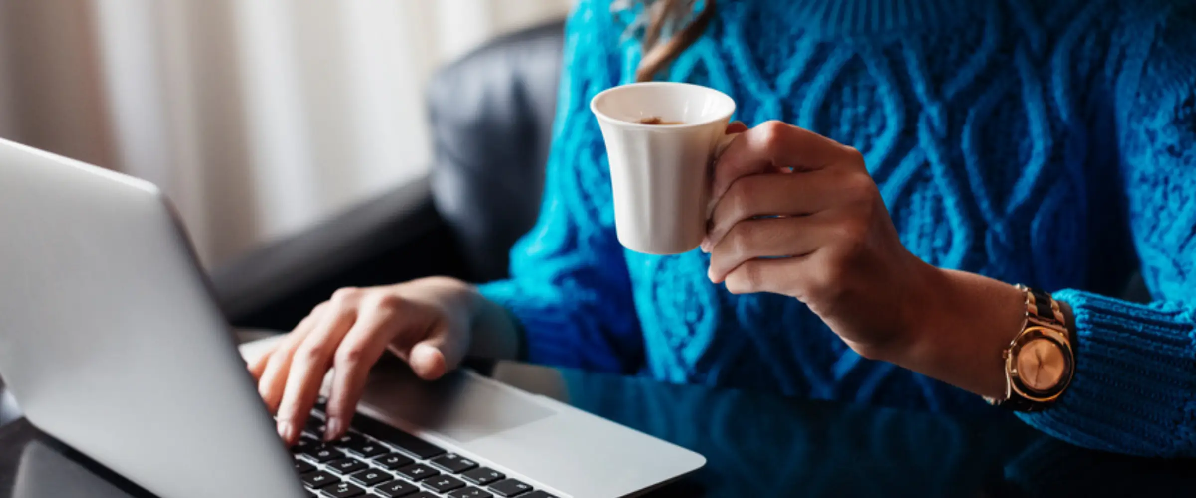 A woman in a blue jumper types on a laptop while holding a cup of espresso coffee.