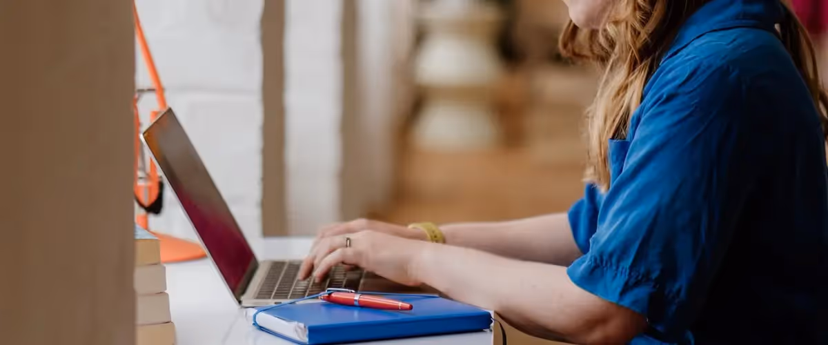 A writer wearing a blue shirt types on a laptop with a notebook and pen beside them.