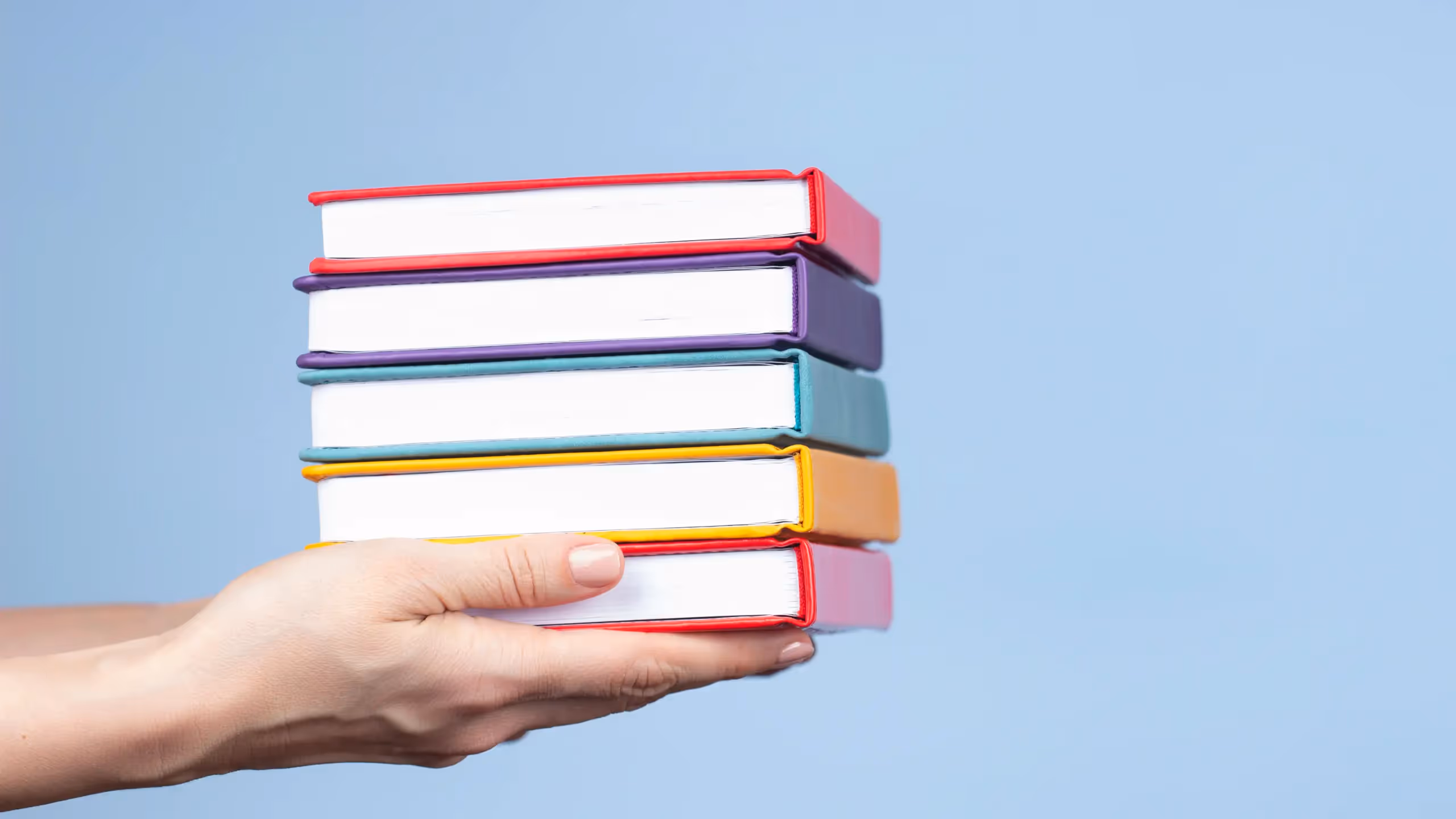 Hands holding out a small stack of hardback books in bright summery colors against a light blue background.