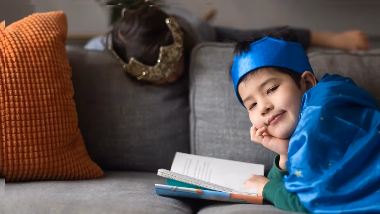 A young boy in a crown with a storybook poses on the sofa while a girl, also in a crown, plays in the background.