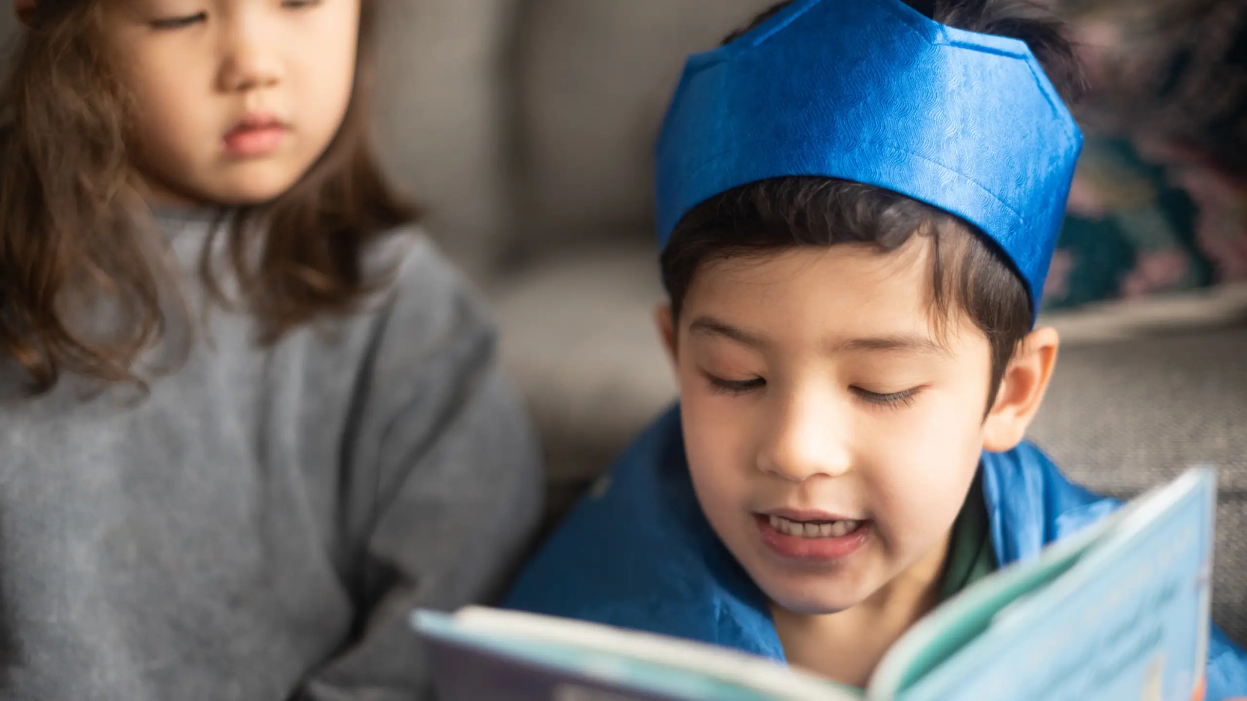 Two young children read a storybook, one of them wearing a blue crown.