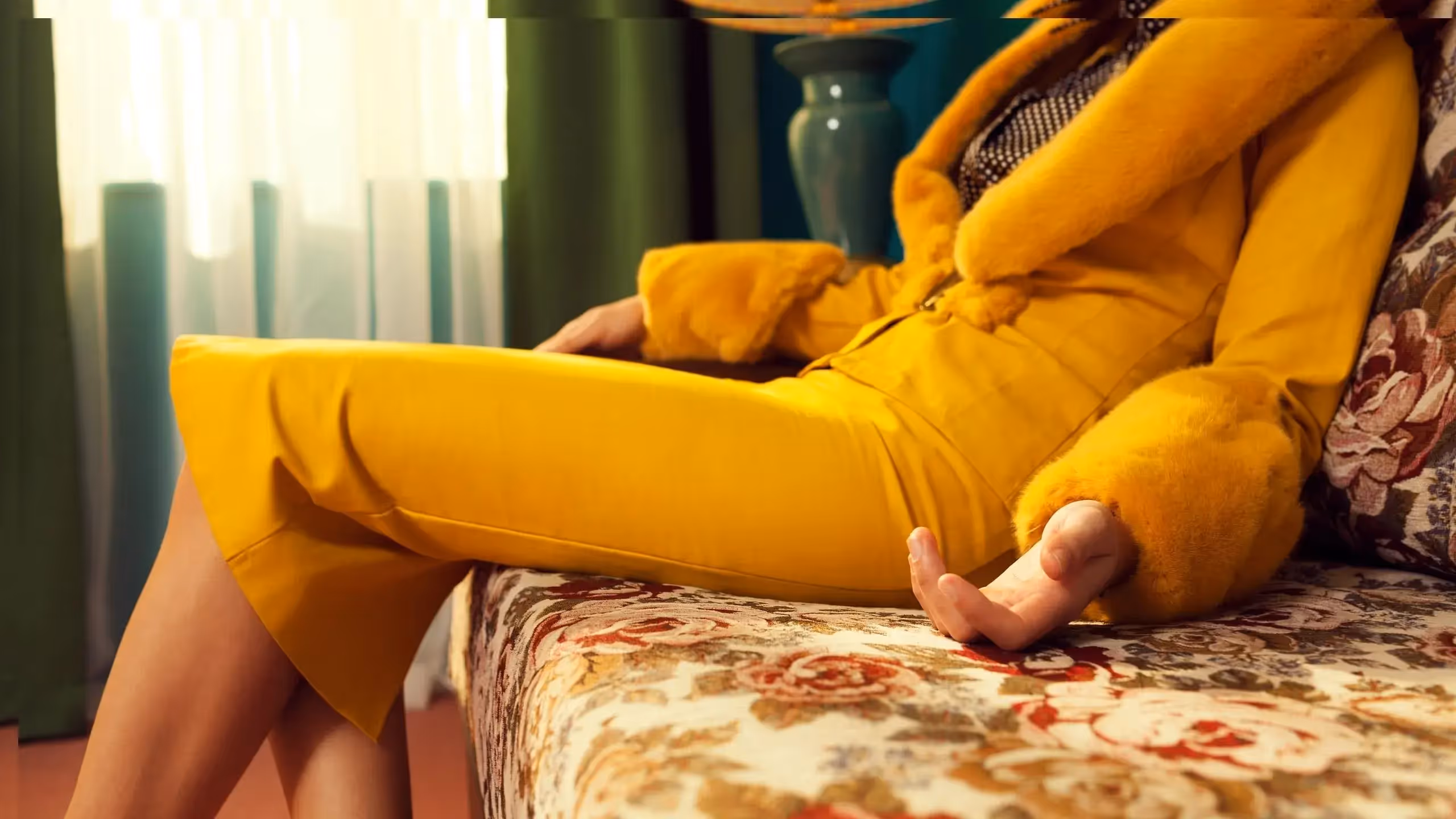 Someone in a yellow outfit sits on a sofa with their arms outstretched. Their head is cut off from the frame.
