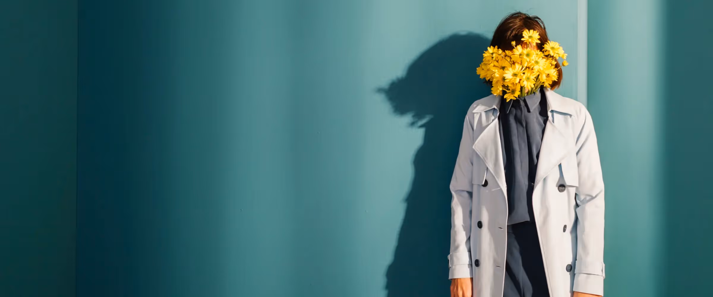 A person in a trenchcoat stands in the corner with flowers covering their face.