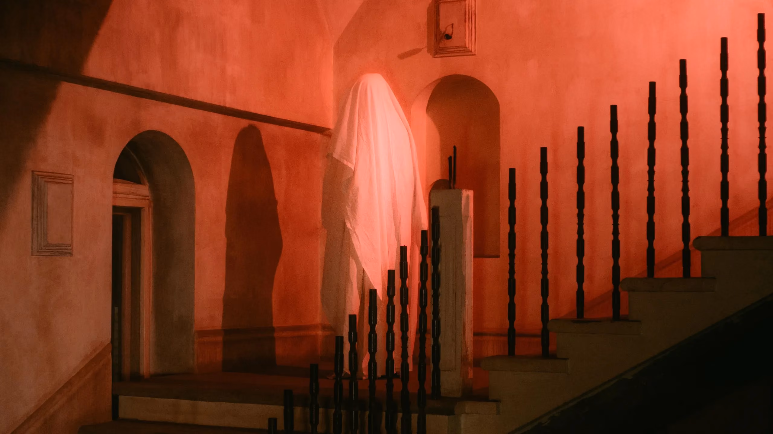 A host drifts up a stairway, the room lit in a mysterious red glow.
