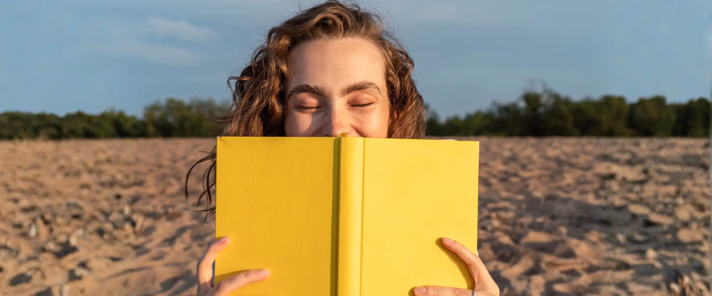 A woman with a yellow book smiles on a beach.