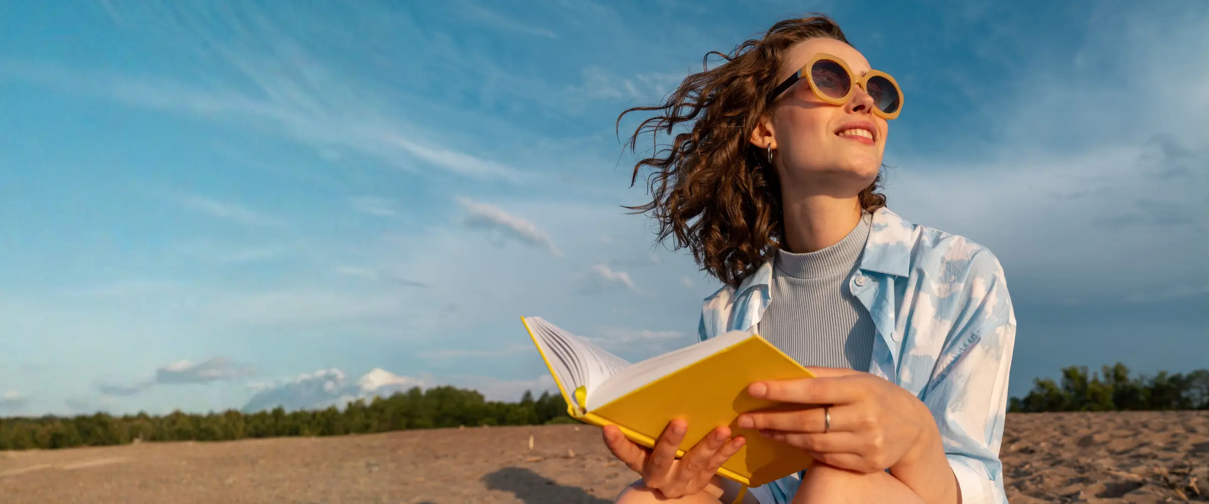 A woman reading a yellow book on a beach with yellow sunglasses.