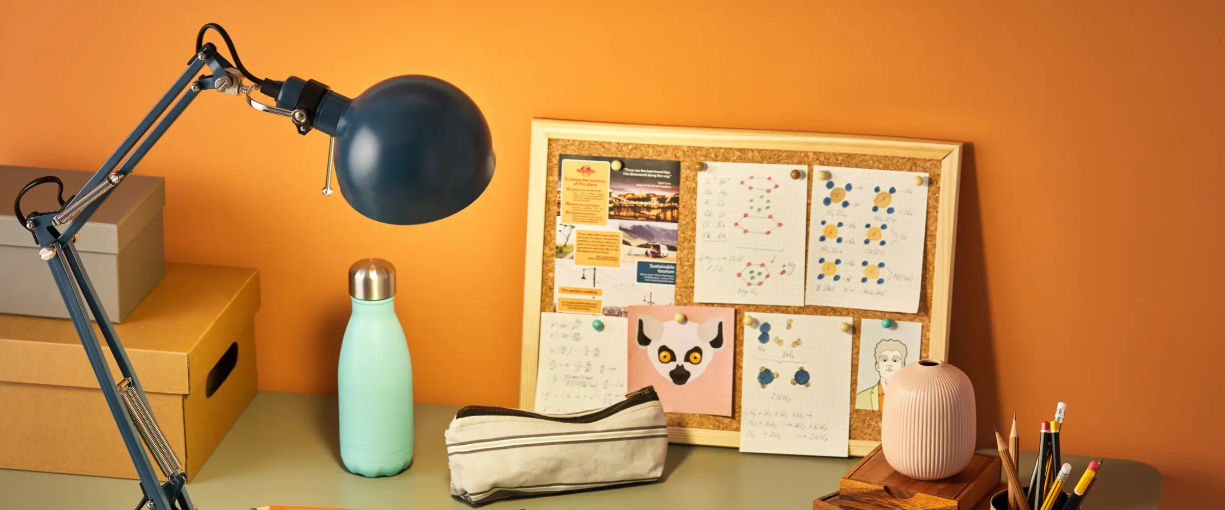 A desk is seen, with a lamp, a pencil case, a water bottle, and a cork board, looking organized.
