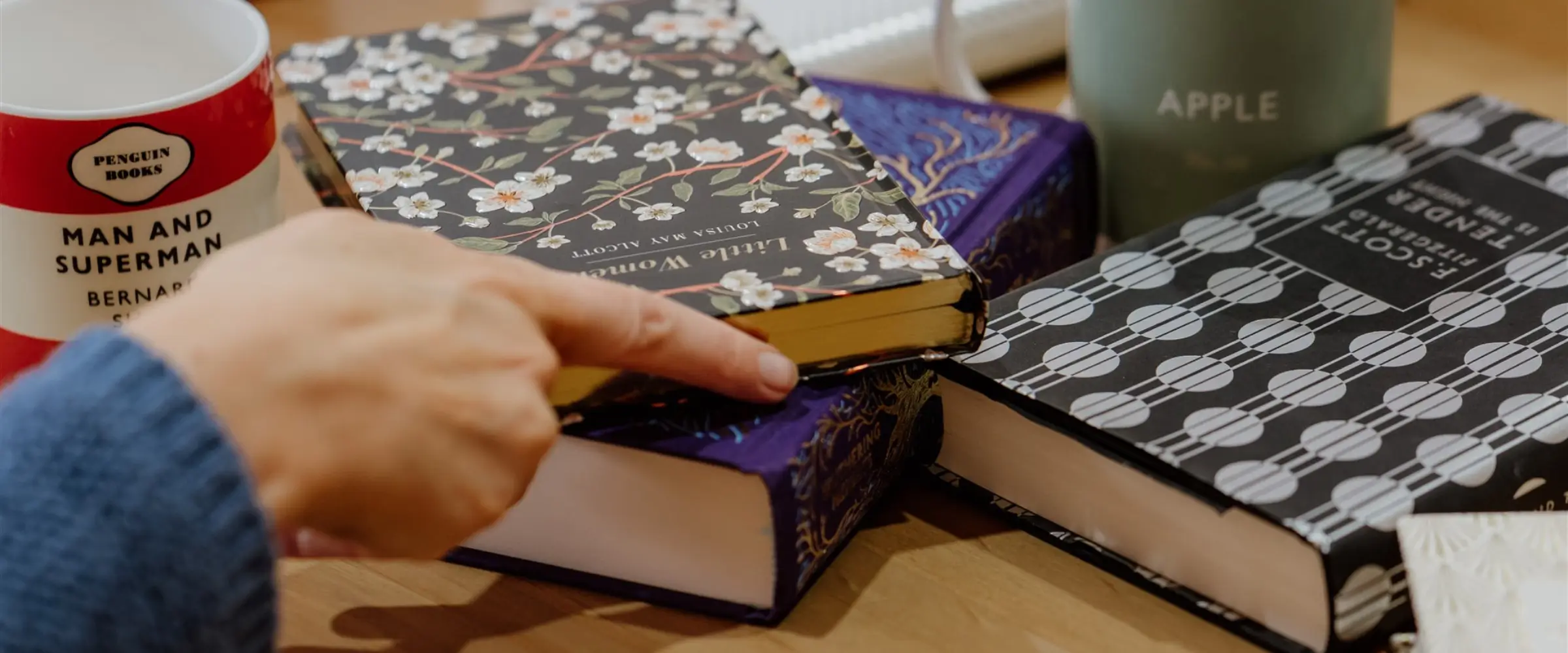 A pile of books sits on a table with mugs, with a hand pointing at them.