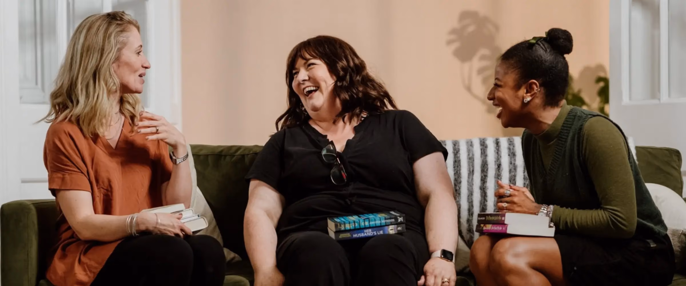 Three women, holding books, sit together on a sofa, laughing.