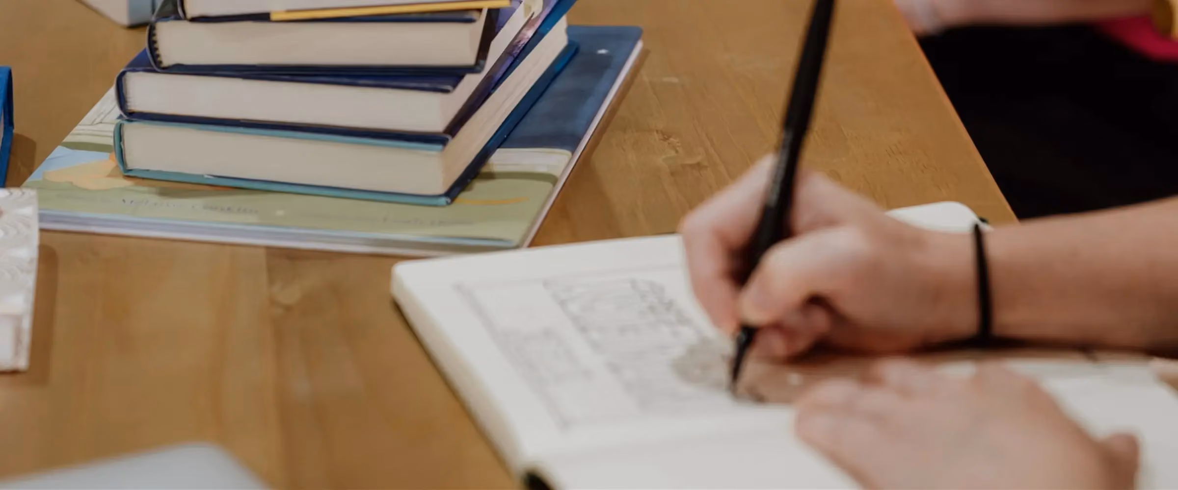 A table is full of books and notebooks. A hand is seen writing in an open book.