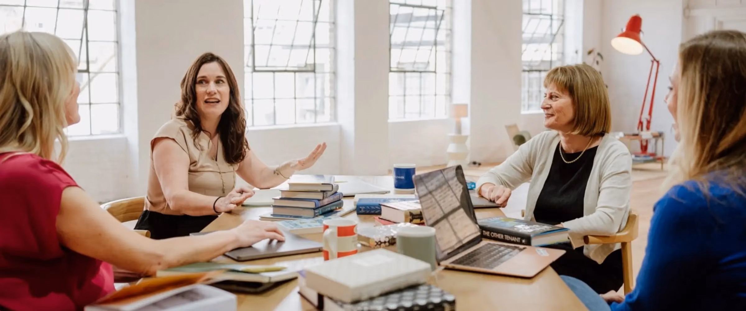 Four female writers sit around a table in conversation, surrounded by books, laptops, and mugs.