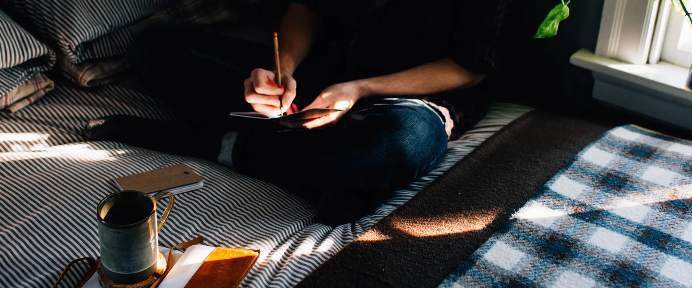 Partial view of a person sitting cross-legged on a bed as they write in a notebook, a mug beside them.