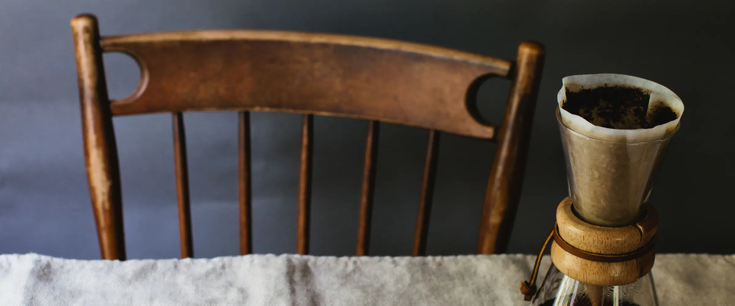 A wooden chair pushed in against a table upon which a coffee-maker sits.