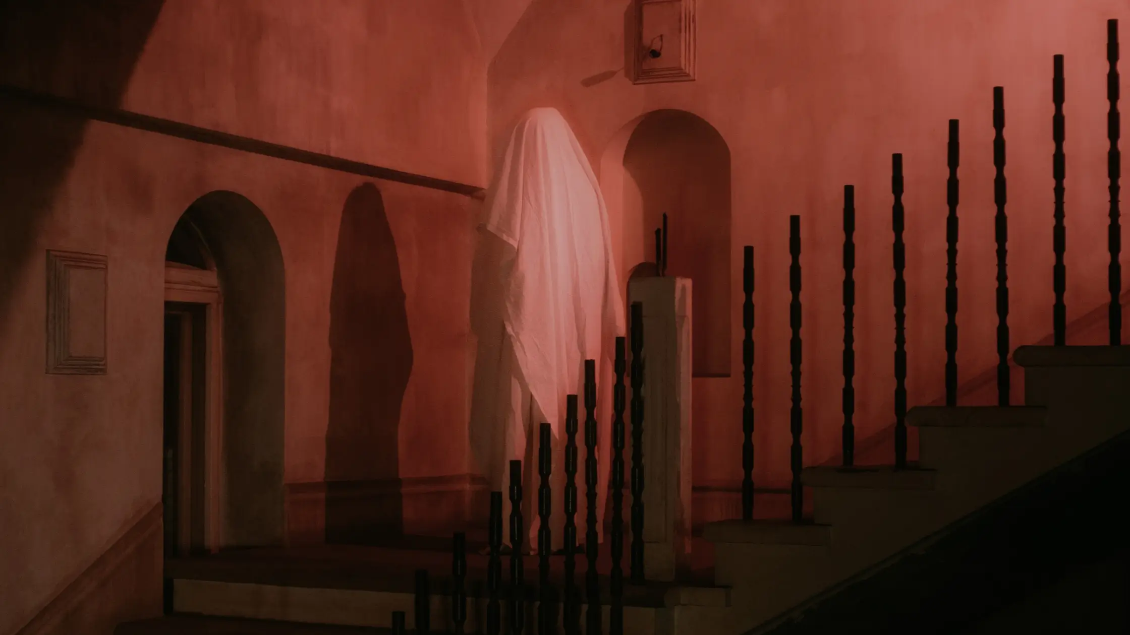 A figure covered in a white sheet stands at the bottom of a red-tinted stairwell.