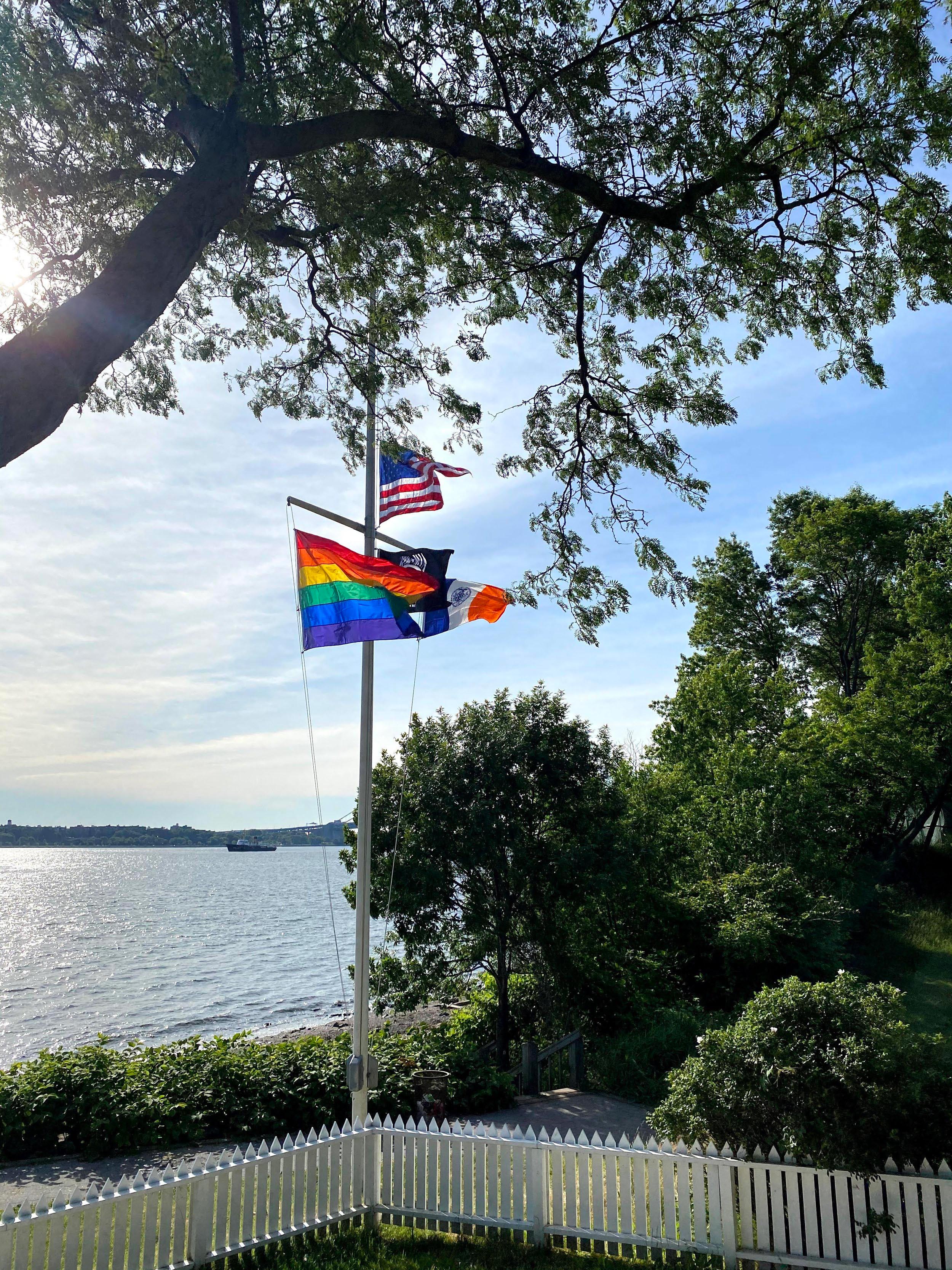 LGBTQ+, USA, and India flags by water with trees and a white fence.