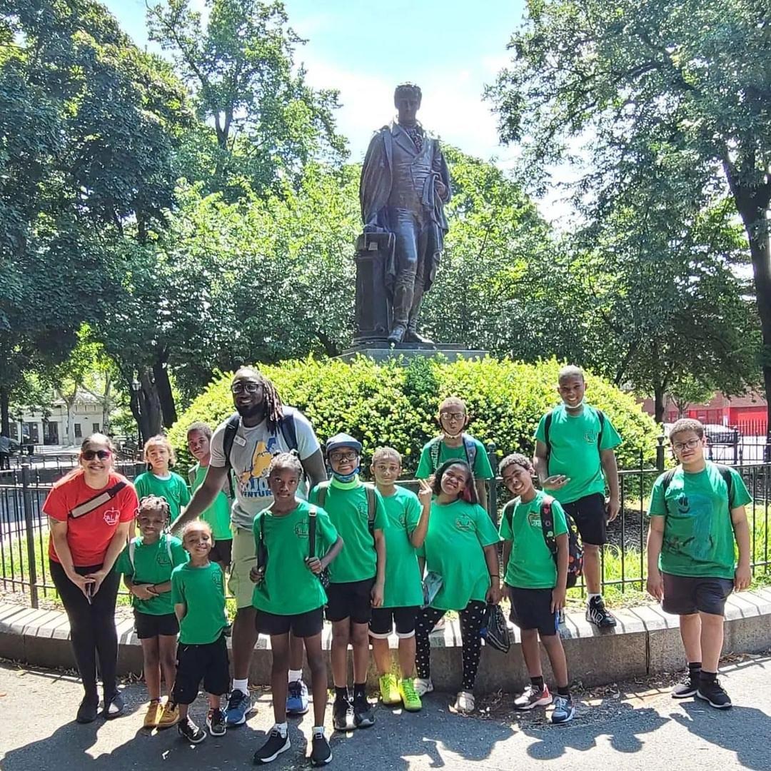 A group of children in green tee shirts pose with their tour guide in front of a bronze statue.