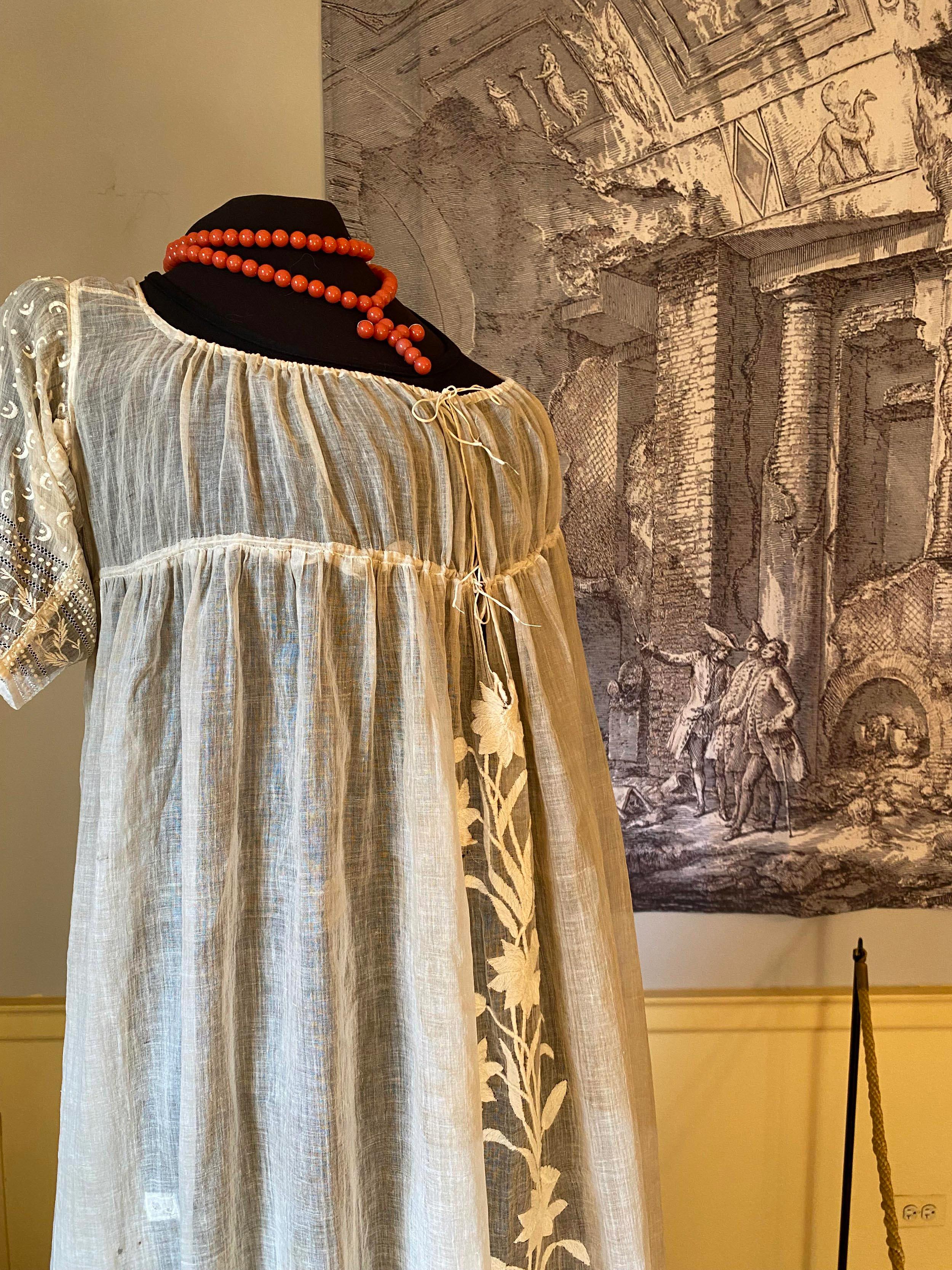 An 1800's style white dress and red rosary are styled on a mannequin.