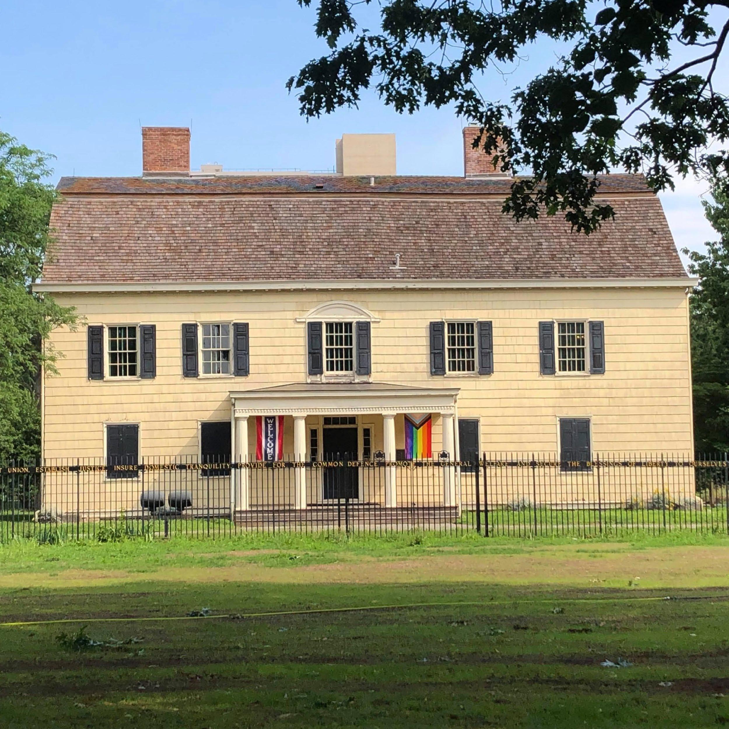 Exterior of King Manor, a yellow house featuring many windows with dark shutters and white columns surrounding the entrance.