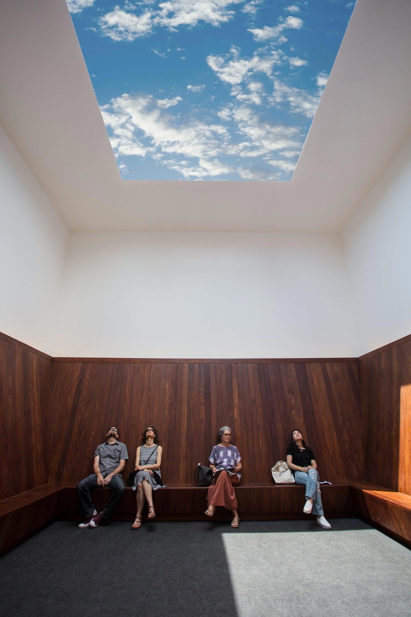 People sit and gaze upward at James Turrell's Meeting, an indoor art installation featuring a unobstructed view of the sky above.