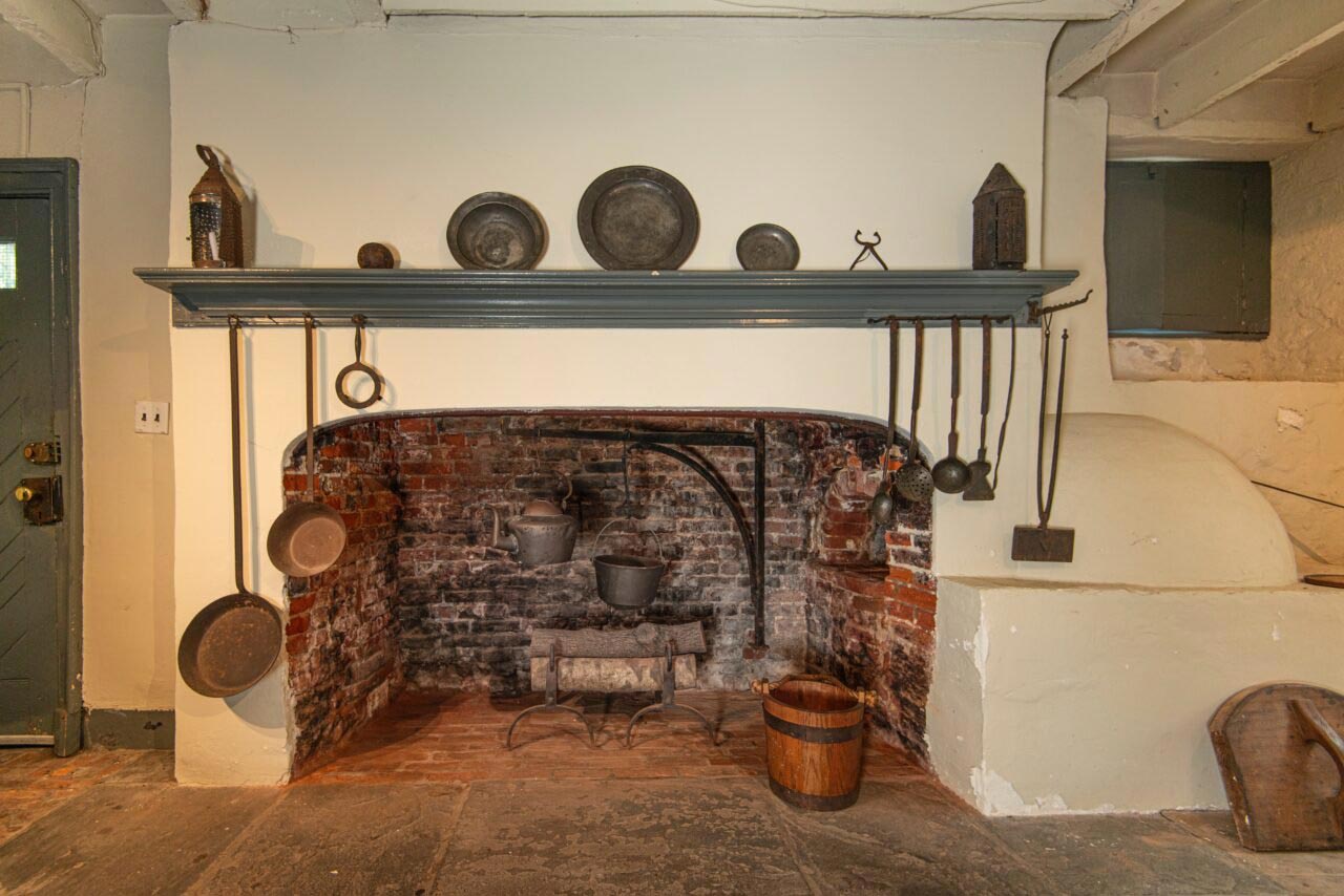 A historic brick hearth is hung round with metal cooking implements and metal plates displayed on a shelf above.