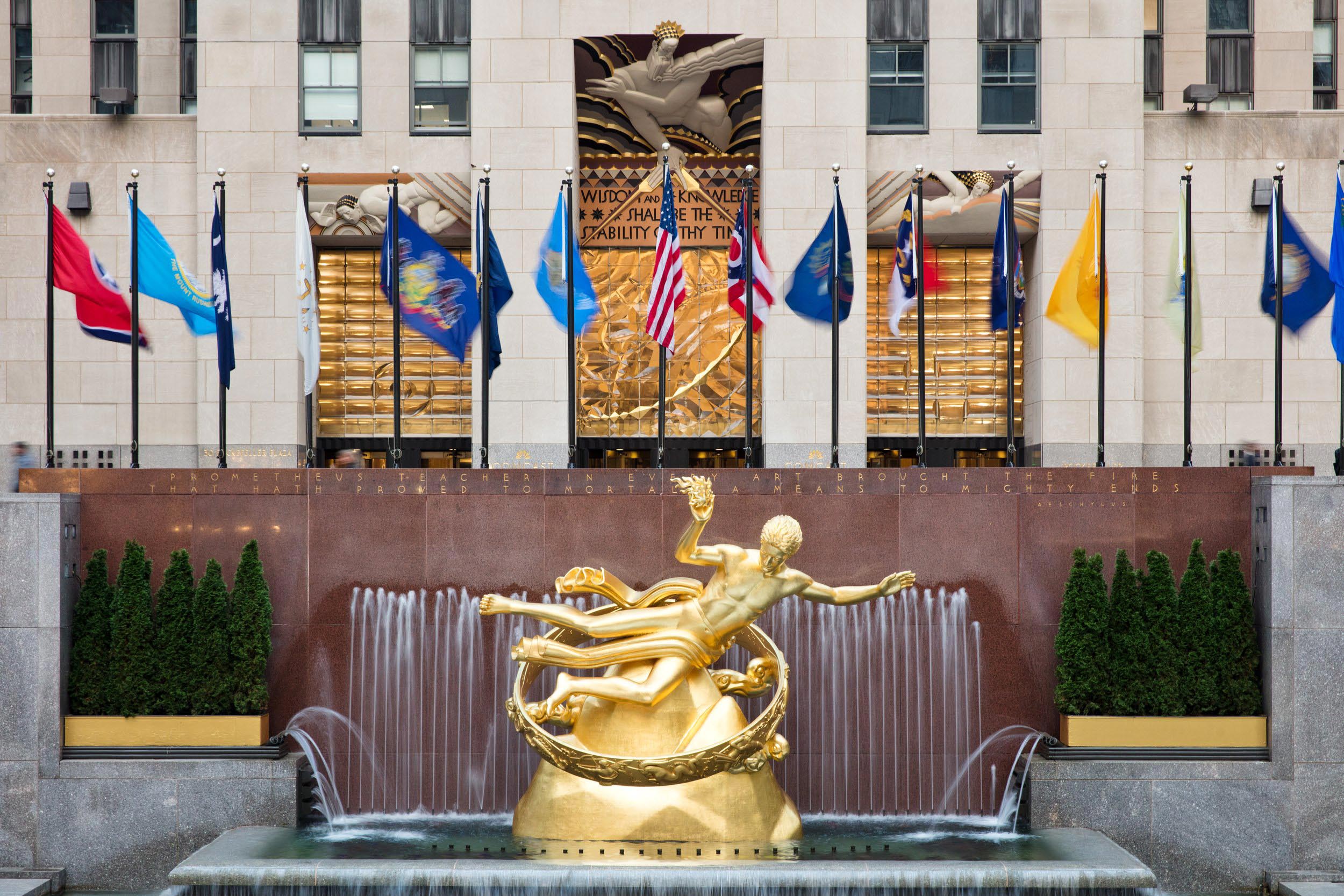 Water cascades around the gilded "Prometheus" fountain while flags of various origins wave in the background.