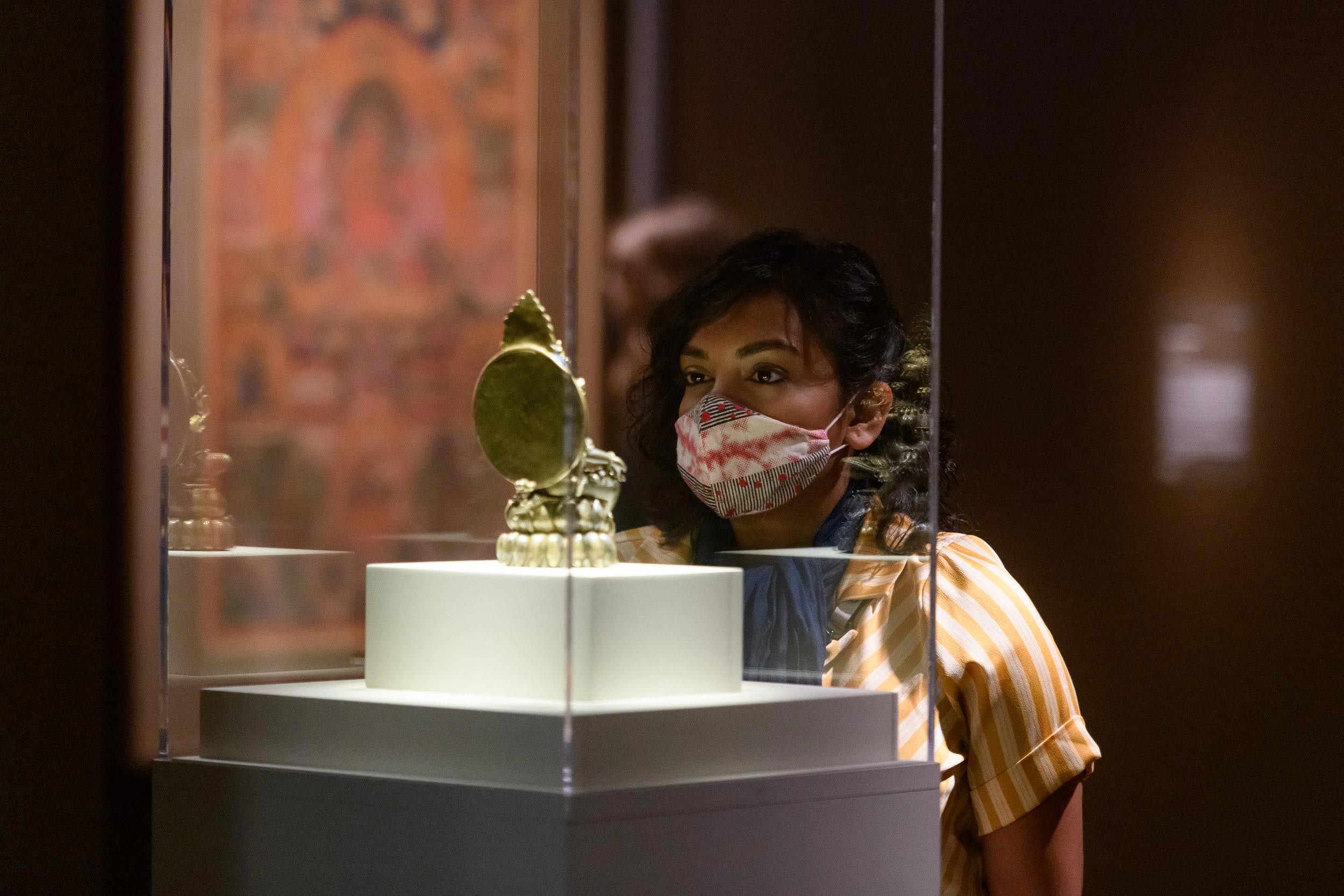 A woman in a mask gazes intently at a small golden artifact in a glass case.