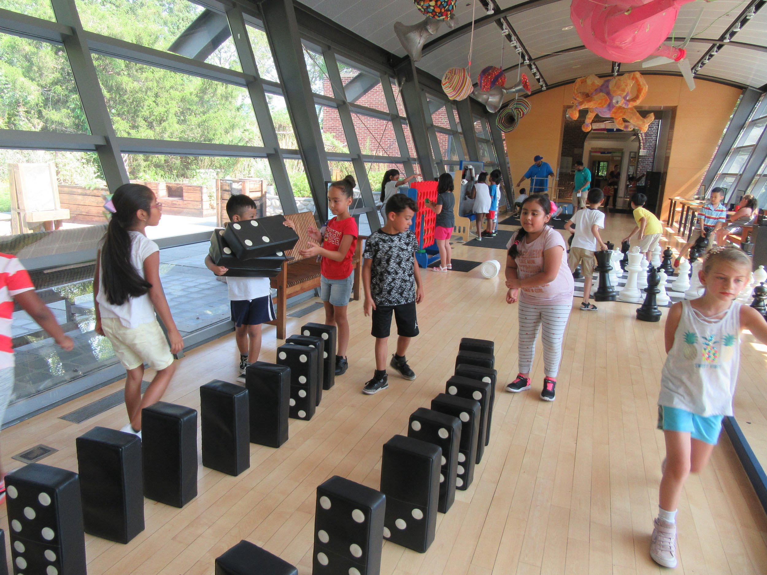 Children play in a room with massively oversized games such as dominos, chess, and Connect 4.