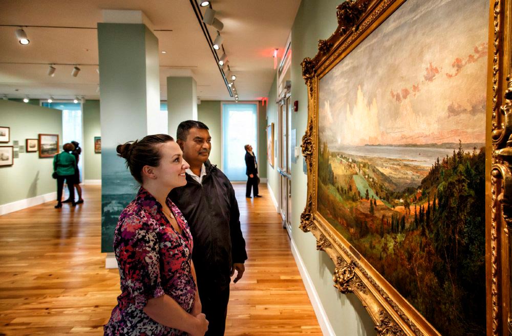 A man and smiling woman gaze at a large painting of a landscape.