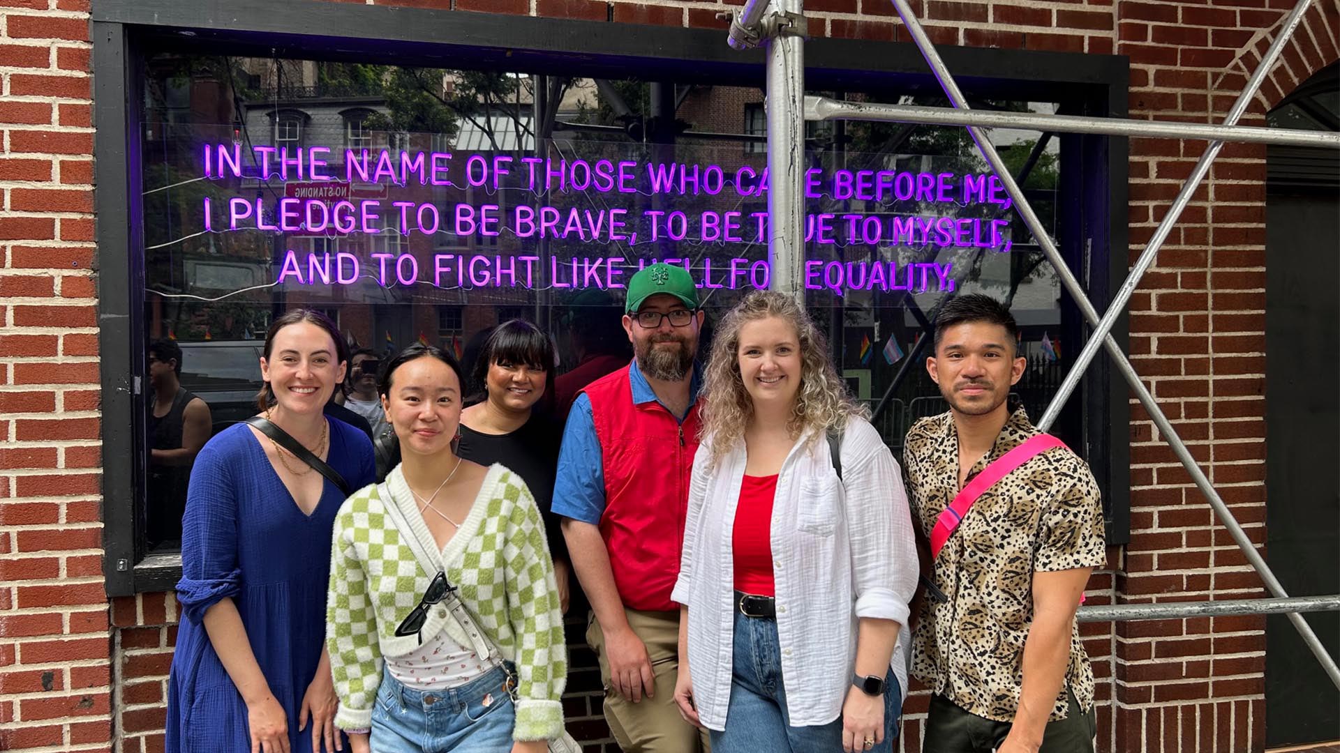 A group of people pose in front of a neon sign stating, "In the name of those who came before me, I pledge to be brave, to be true to myself, and to fight like hell for equality."