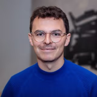 Smiling man with glasses and short curly hair wearing a blue sweater against a blurred background.