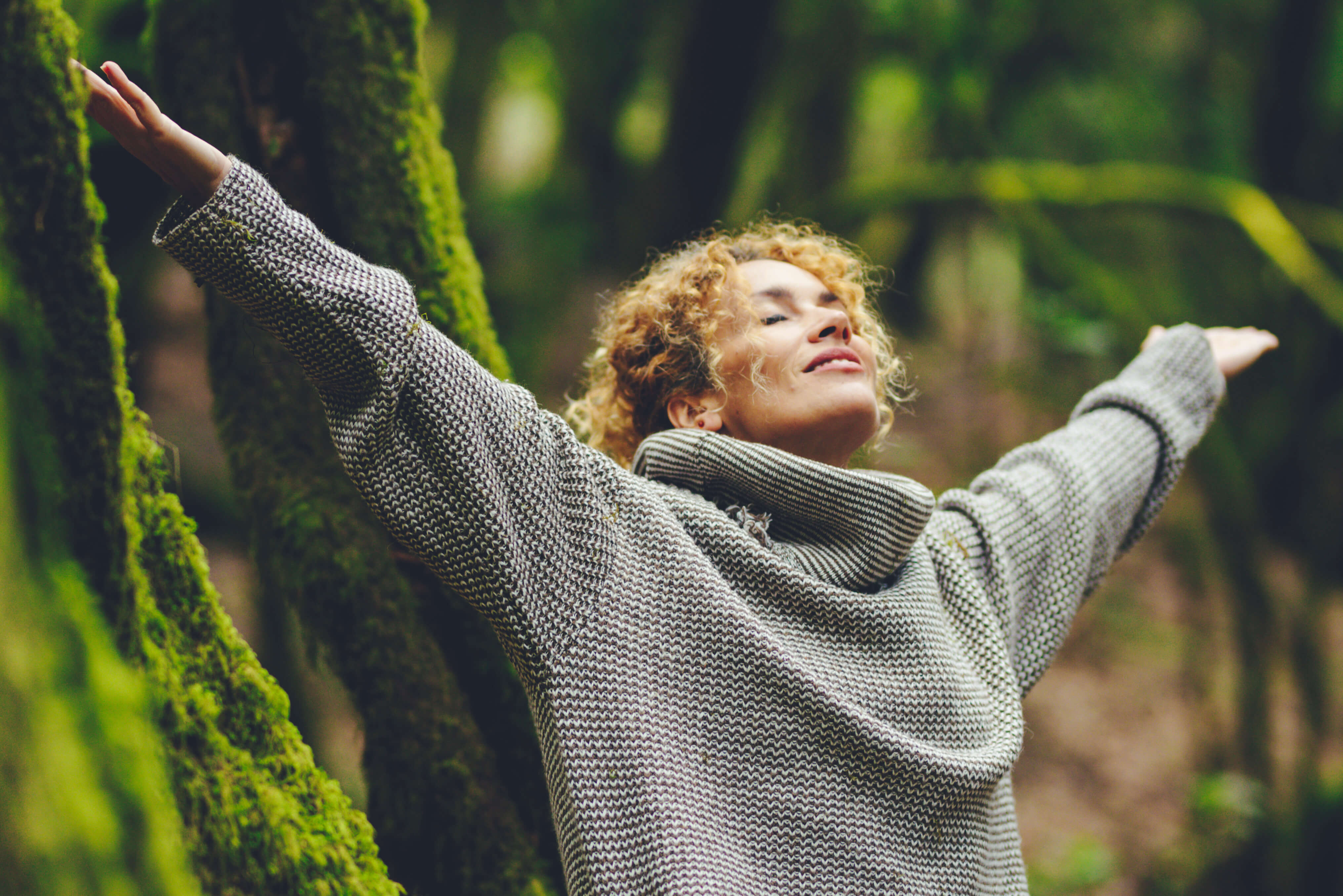 Woman with curly hair and grey kit sweater in nature throwing her hands in the air. 