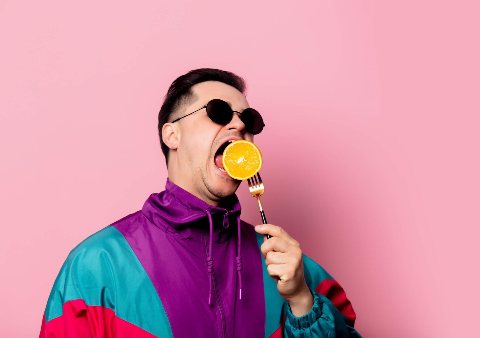 homme avec lunettes de soleil mangeant une orange sur une fourchette sur fond rose clair