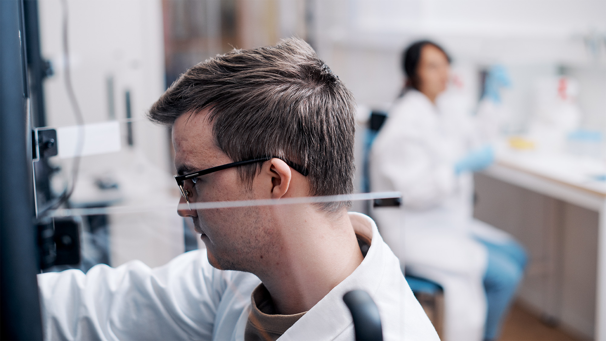 Close-up of a researcher in lab coat, shown in profile. The researcher is reaching into a scientific instrument that is located outside of the picture. The researcher's face is partly obscured by the opened transparent door of the instrument.