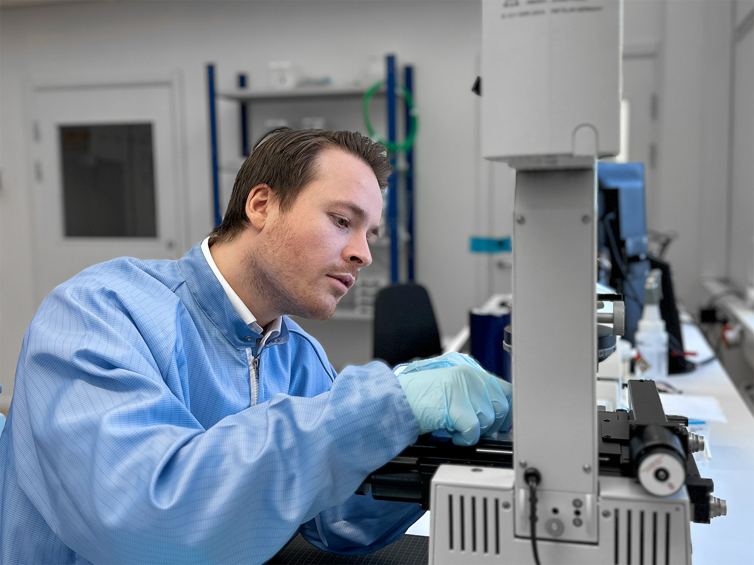 Person in a blue lag coat and green-blue nitrile gloves sitting in front of a microscope. The image shows the top half of the persons body. His hands is raised to manipulate an object on the micoscope and he is looking down on his hands with intense focus.