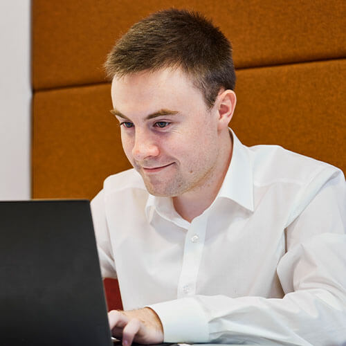 Johannes Heeger in white shirt working on a laptop in an office with brown padded walls.