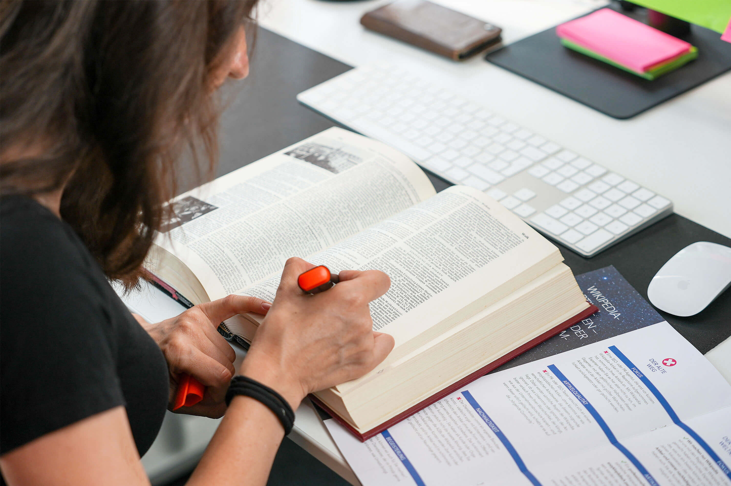 Person highlighting text in an open book at a desk with a keyboard, mouse, and documents nearby.
