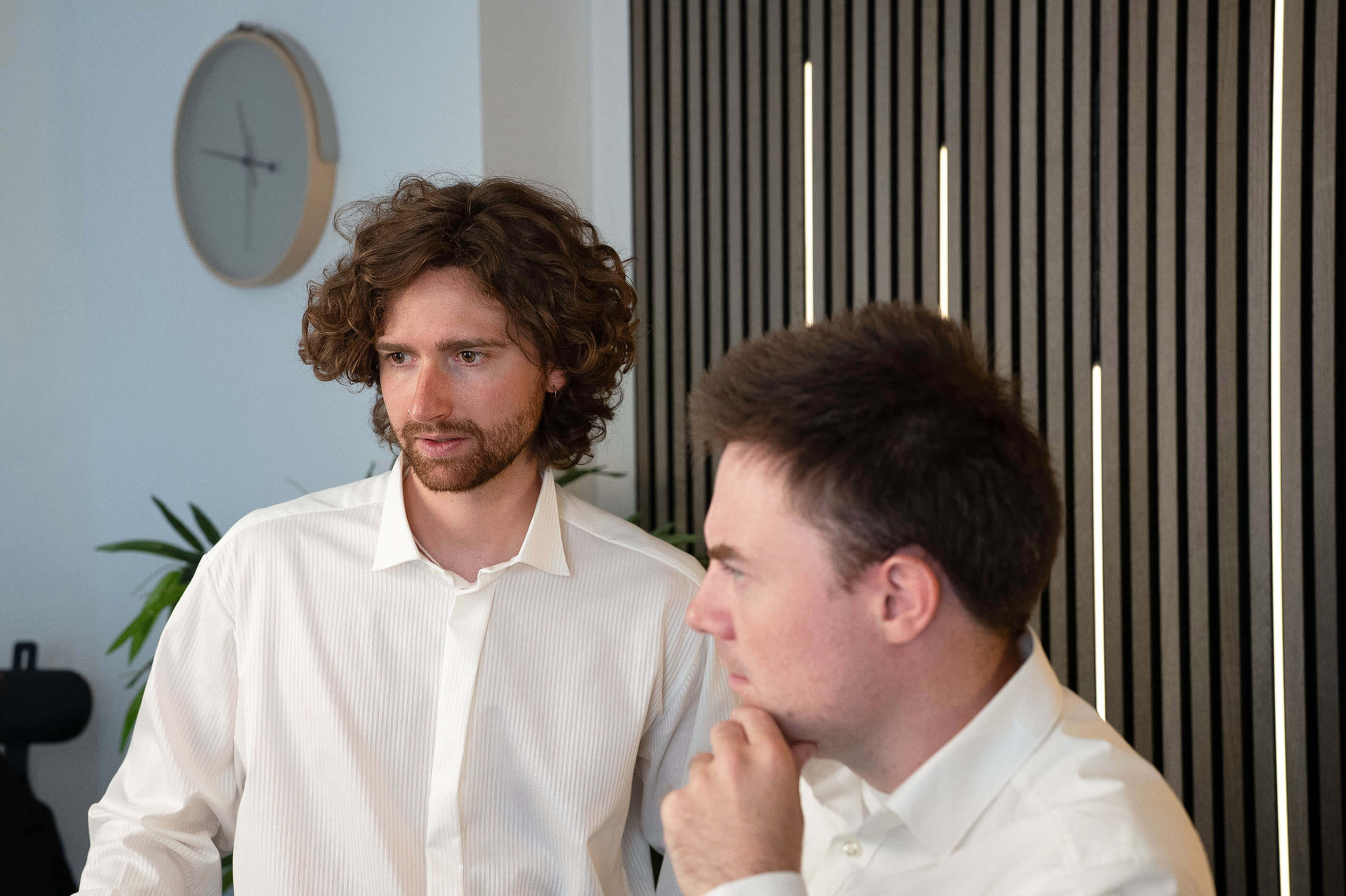 Two men in white shirts engaged in discussion in an office setting with a wall clock and vertical blinds in the background.