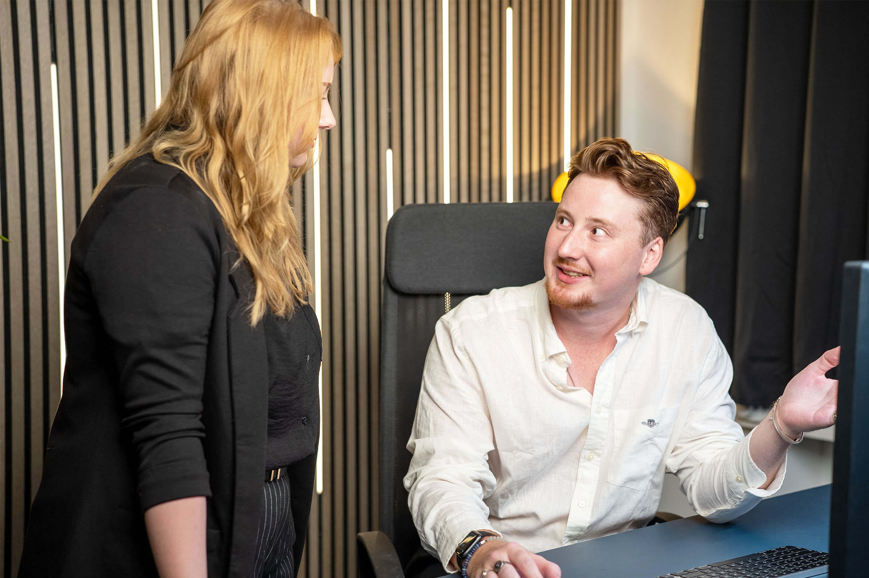 Man in white shirt sitting at a desk talking to a standing woman with blonde hair and black blazer.