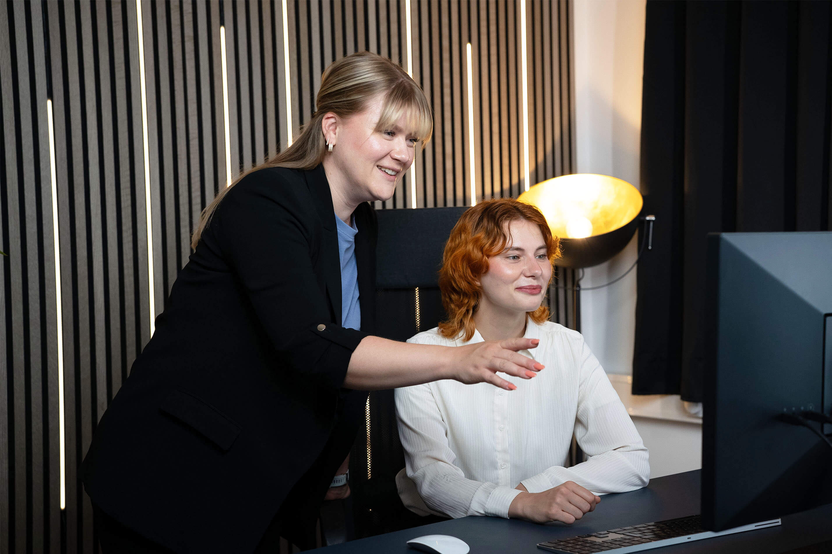Two women collaborating at a desk, one pointing at a computer screen while the other watches attentively.