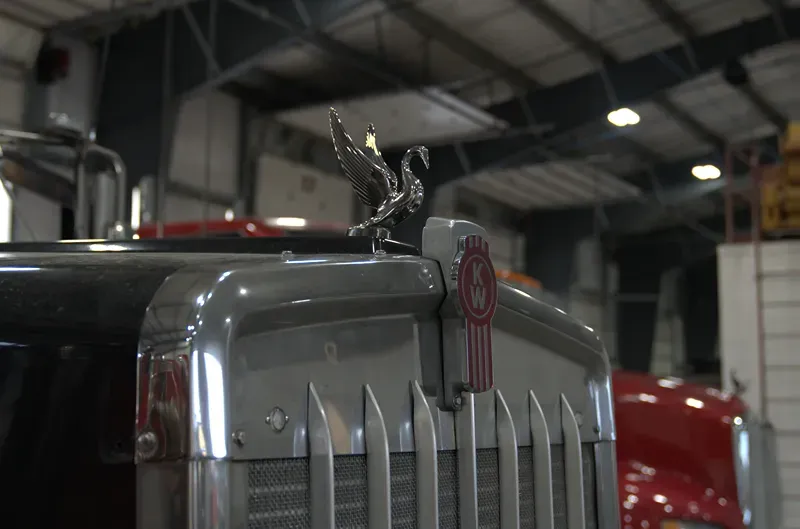 Heavy-duty Kenworth truck grille with red “KW” badge and chrome swan hood ornament inside a repair shop.