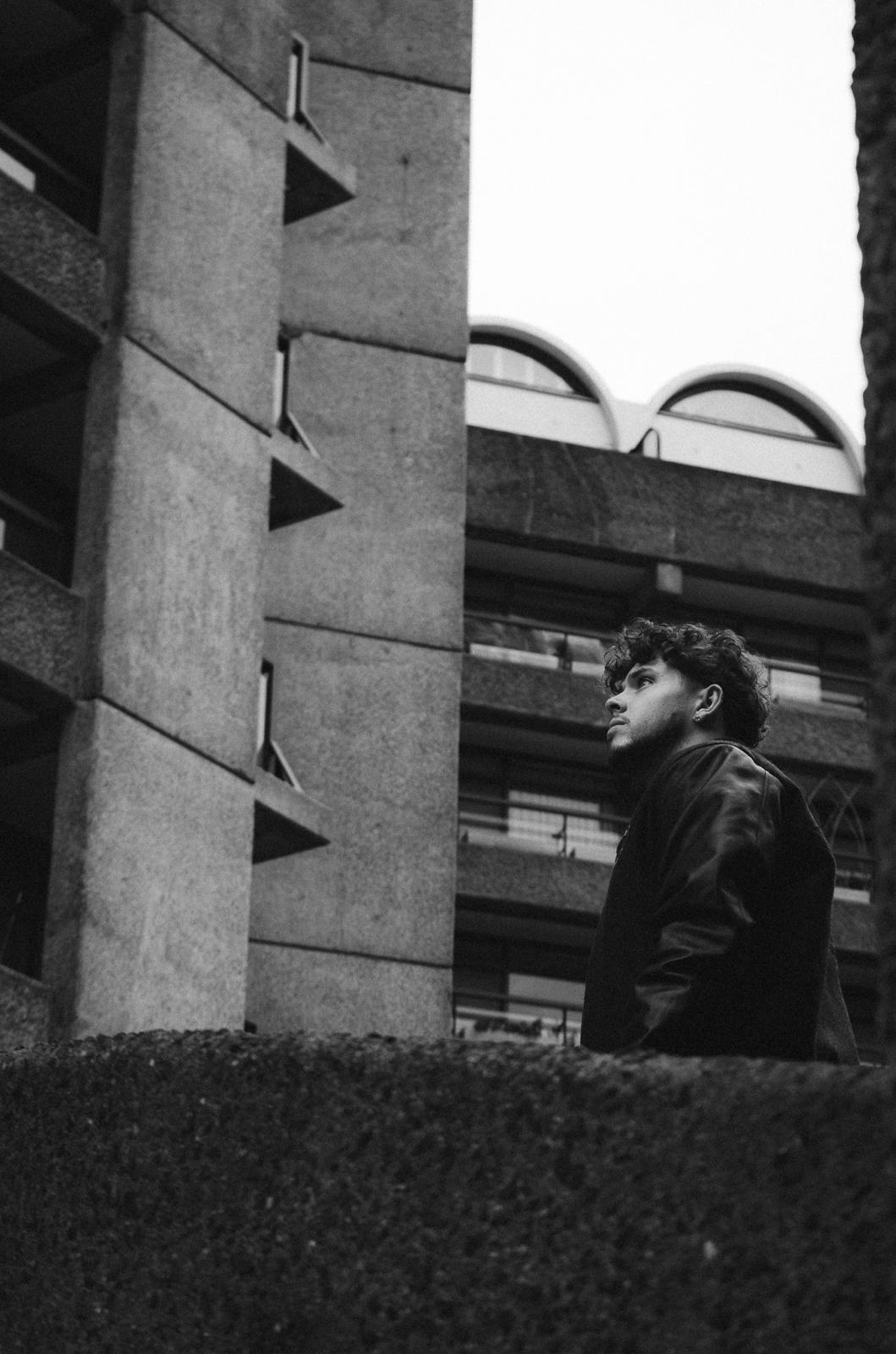 Black and white photo of a man with curly hair in a jacket looking up between brutalist concrete buildings.