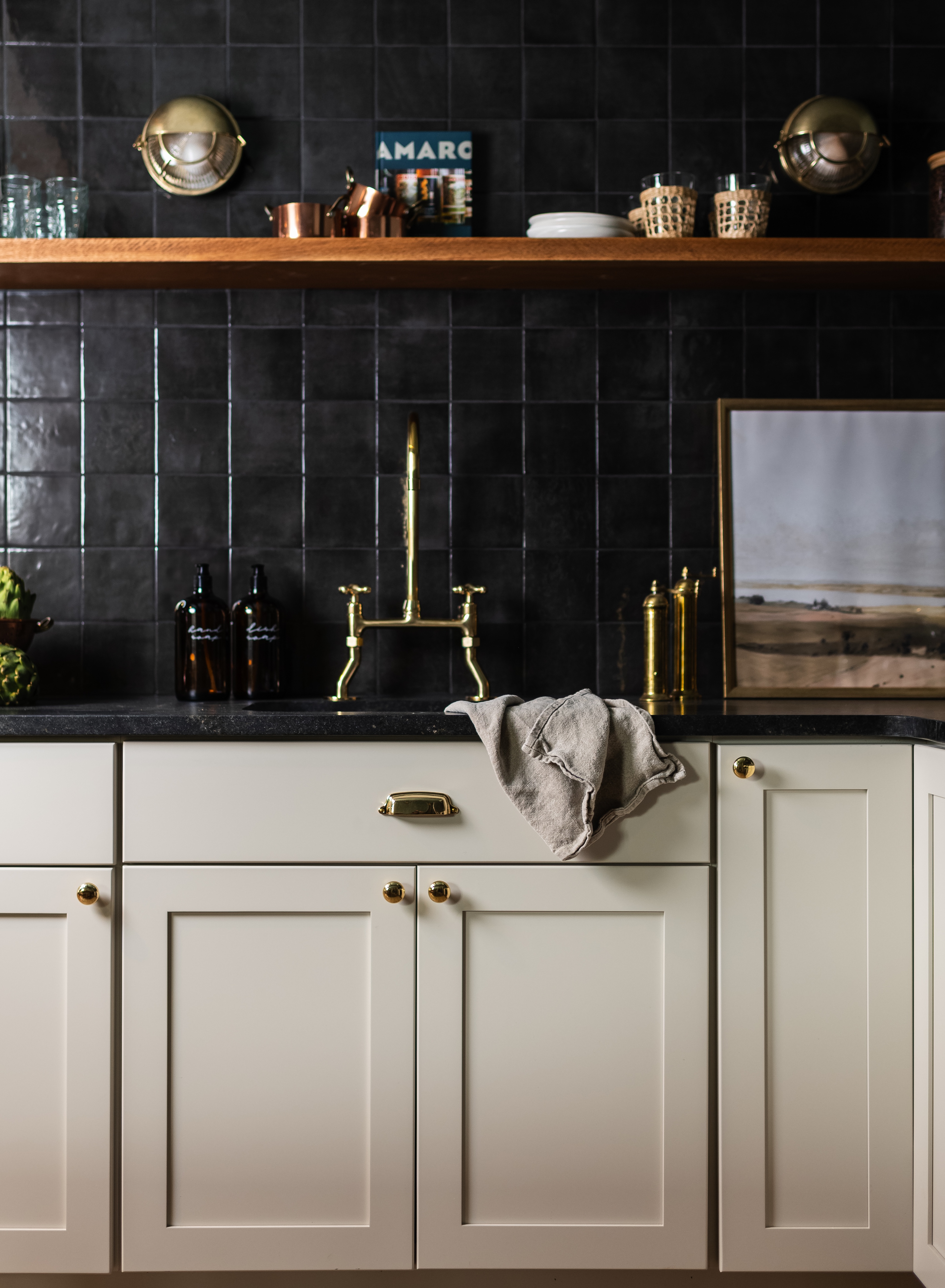 Modern kitchen sink area with brass faucet, beige cabinets, black tile backsplash, wooden shelf with dishes and decor.