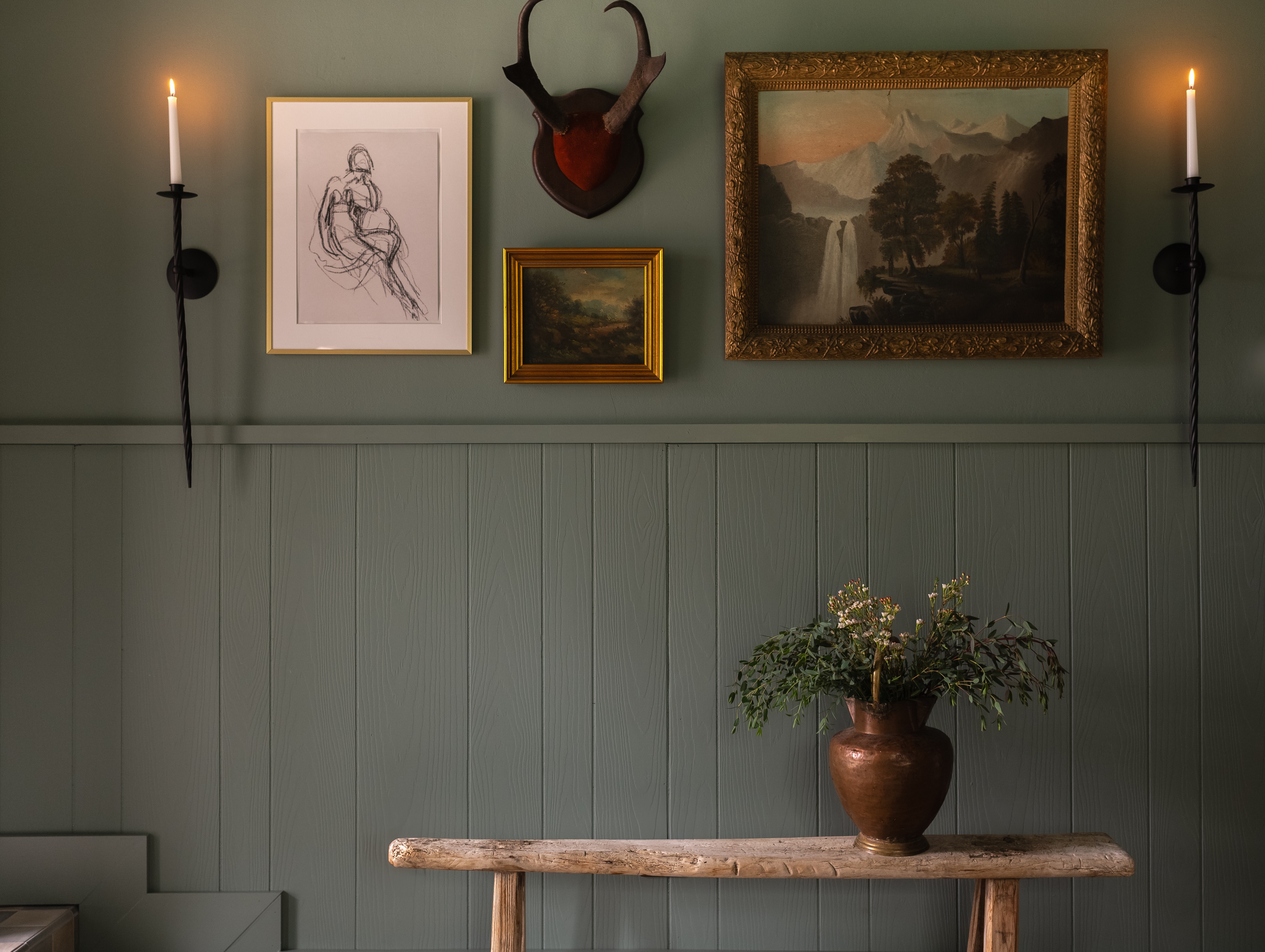 Rustic interior wall with green wainscoting, two lit wall candles, framed artwork, mounted antlers, and a wooden bench holding a copper vase with greenery.