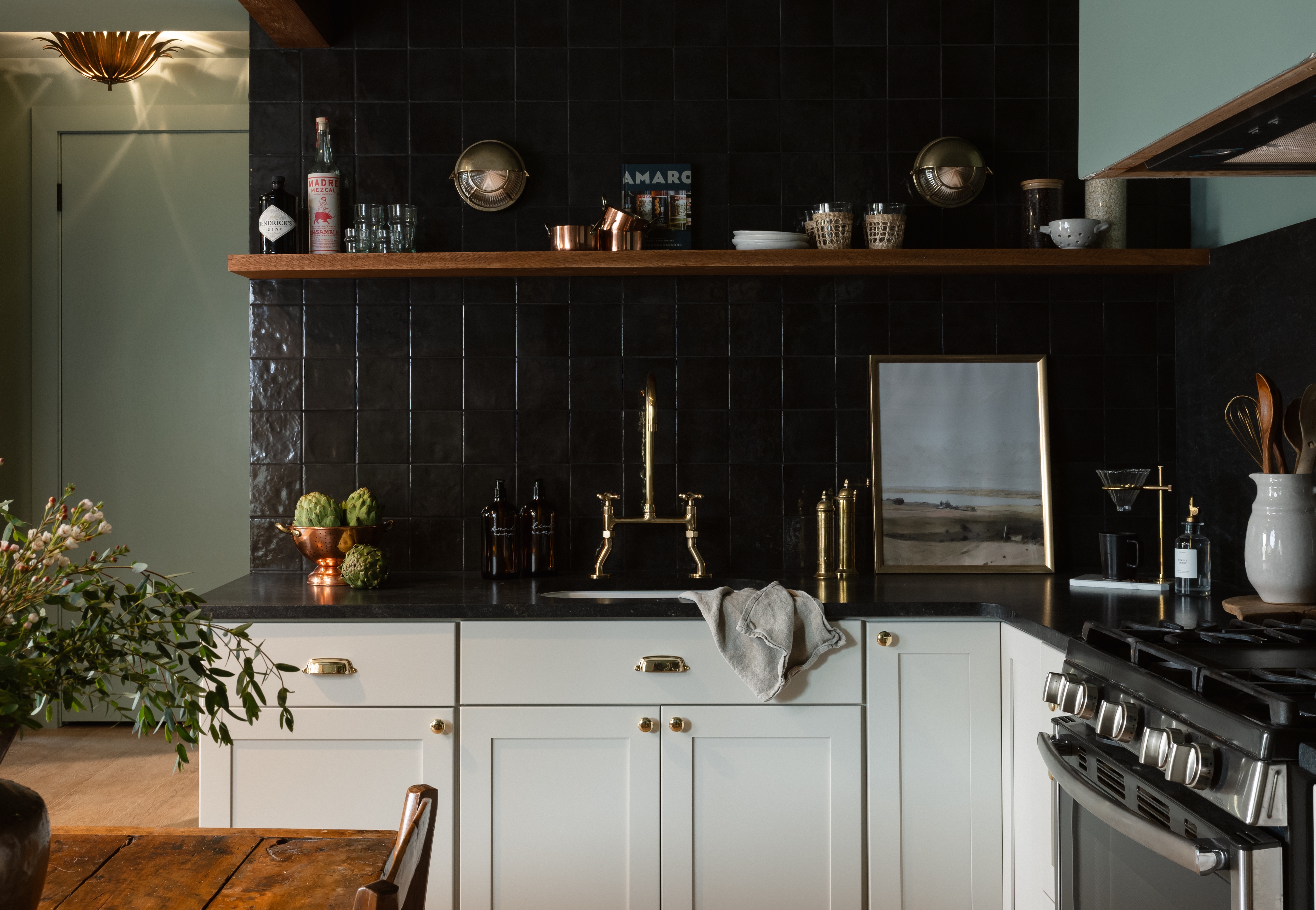 Modern kitchen with white cabinets, black tiled backsplash, brass faucet, wooden shelf holding glassware, and a stove on the right.