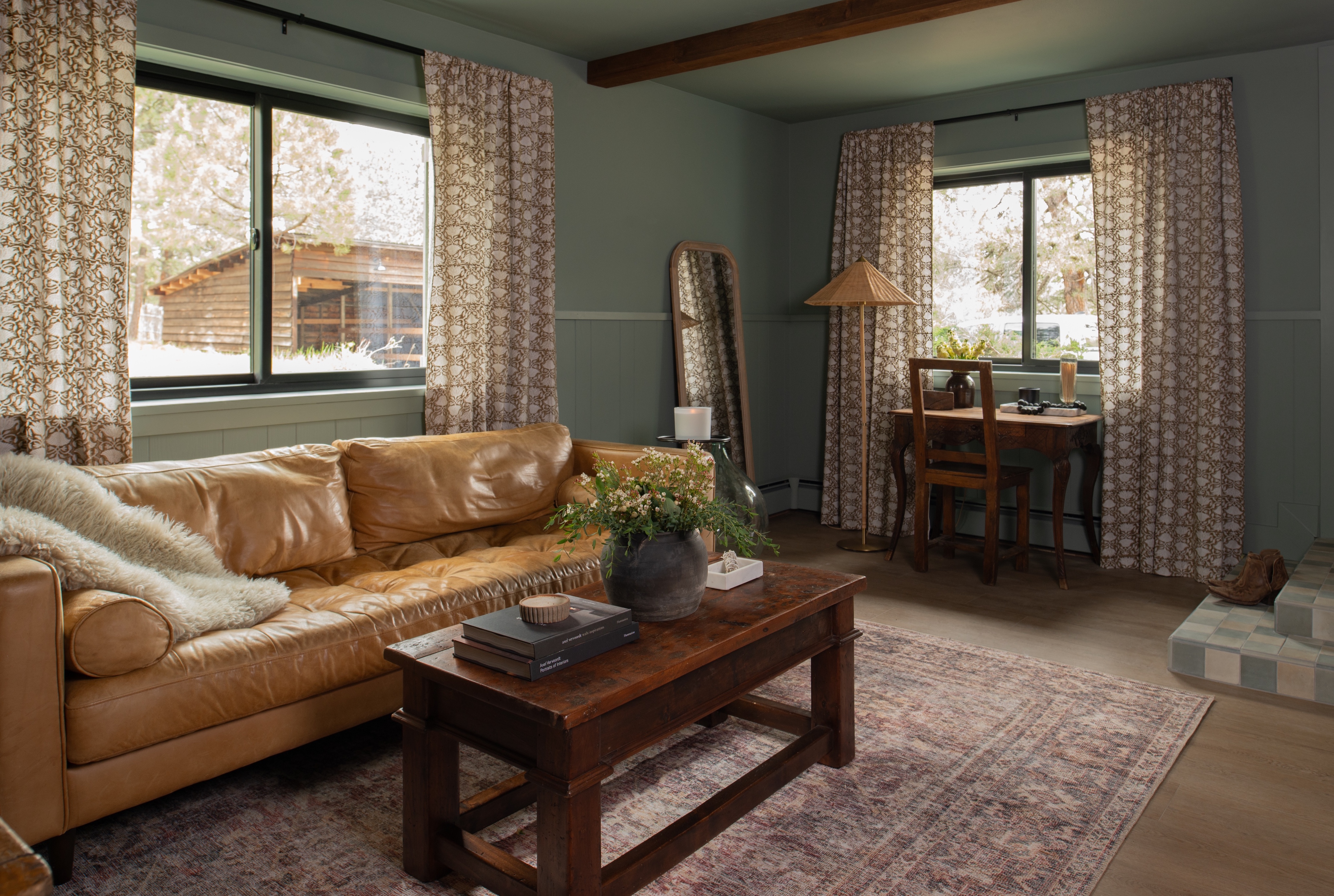 Cozy living room with tan leather sofa, patterned curtains, wooden coffee table with books and a plant, floor mirror, and a small wooden desk with a chair.