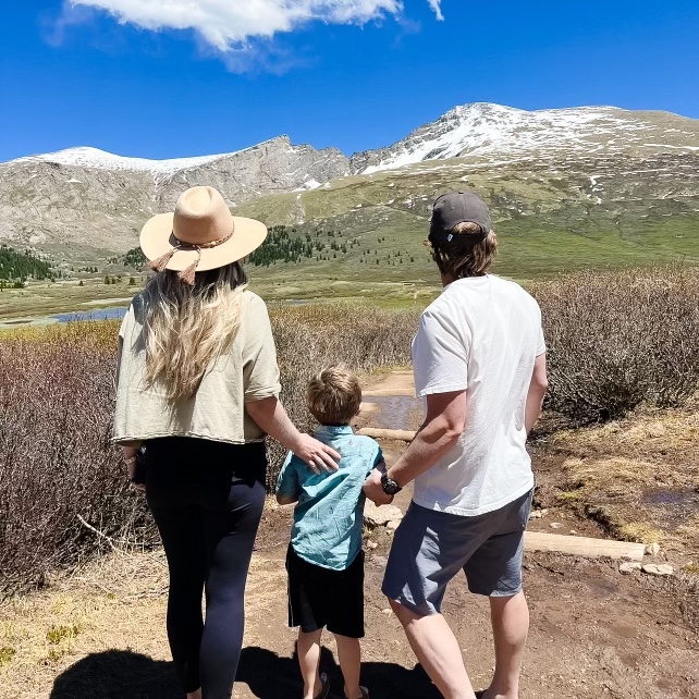 A family of three holding hands and looking at snow-capped mountains under a blue sky.