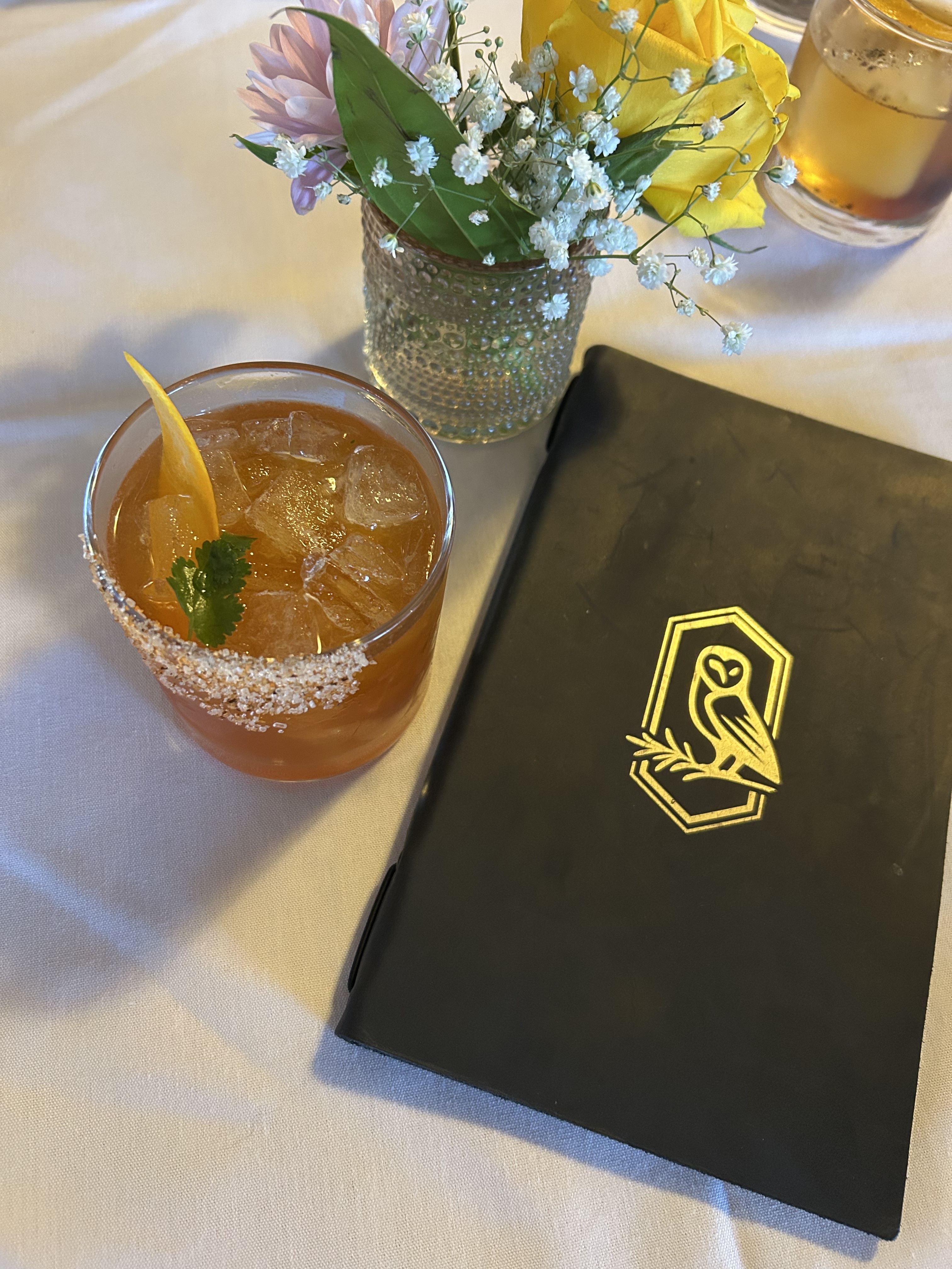Orange cocktail with ice, an orange peel, and a herb garnish next to a black menu with a gold owl emblem, and a small floral arrangement on a white tablecloth.