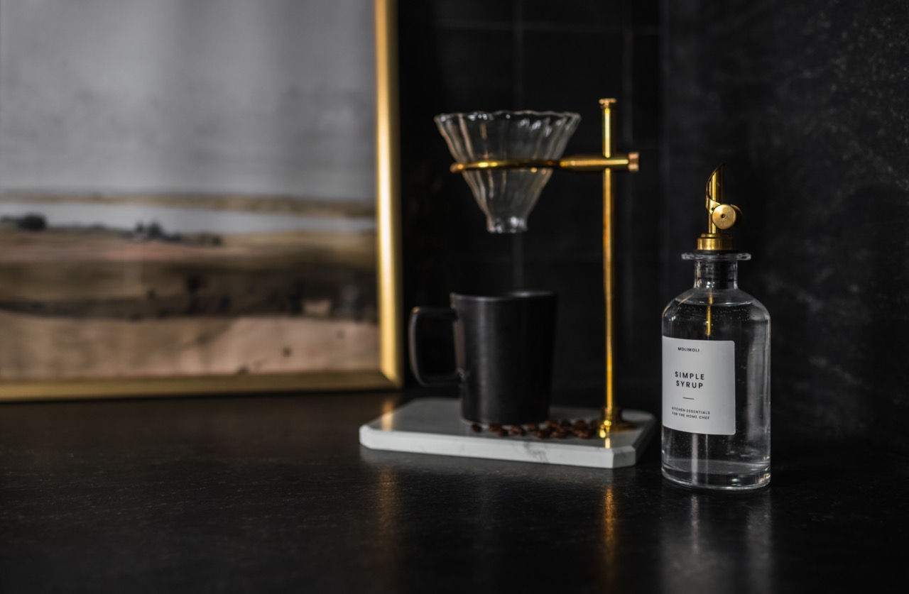 Black countertop with a glass bottle labeled 'Simple Syrup' and a pour-over coffee maker with a black mug and coffee beans.
