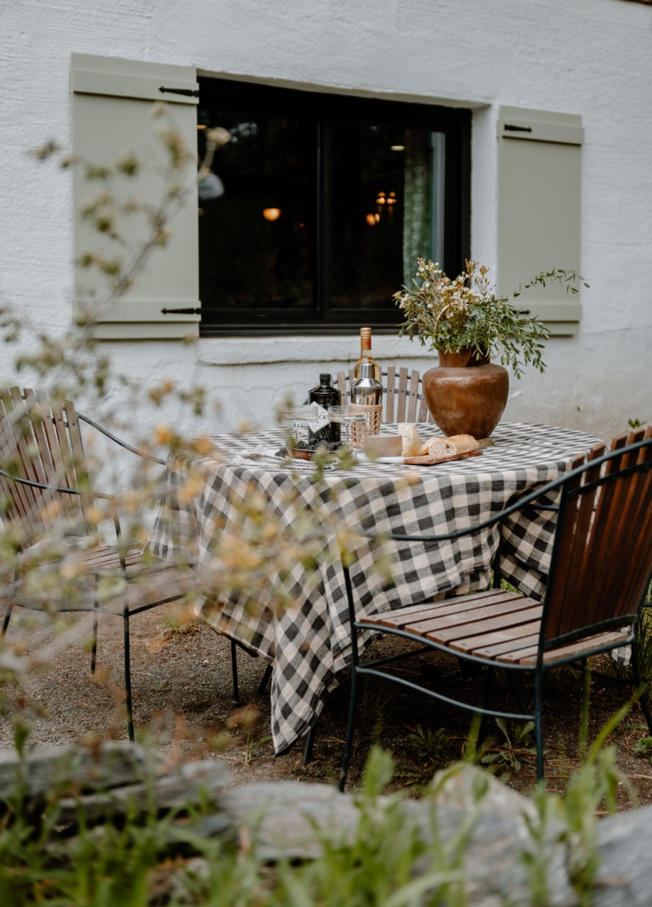 Outdoor wooden table covered with black-and-white checkered tablecloth, set with bottles, glasses, bread, cheese, and a brown vase with flowers, surrounded by wooden chairs near a white wall with a window.